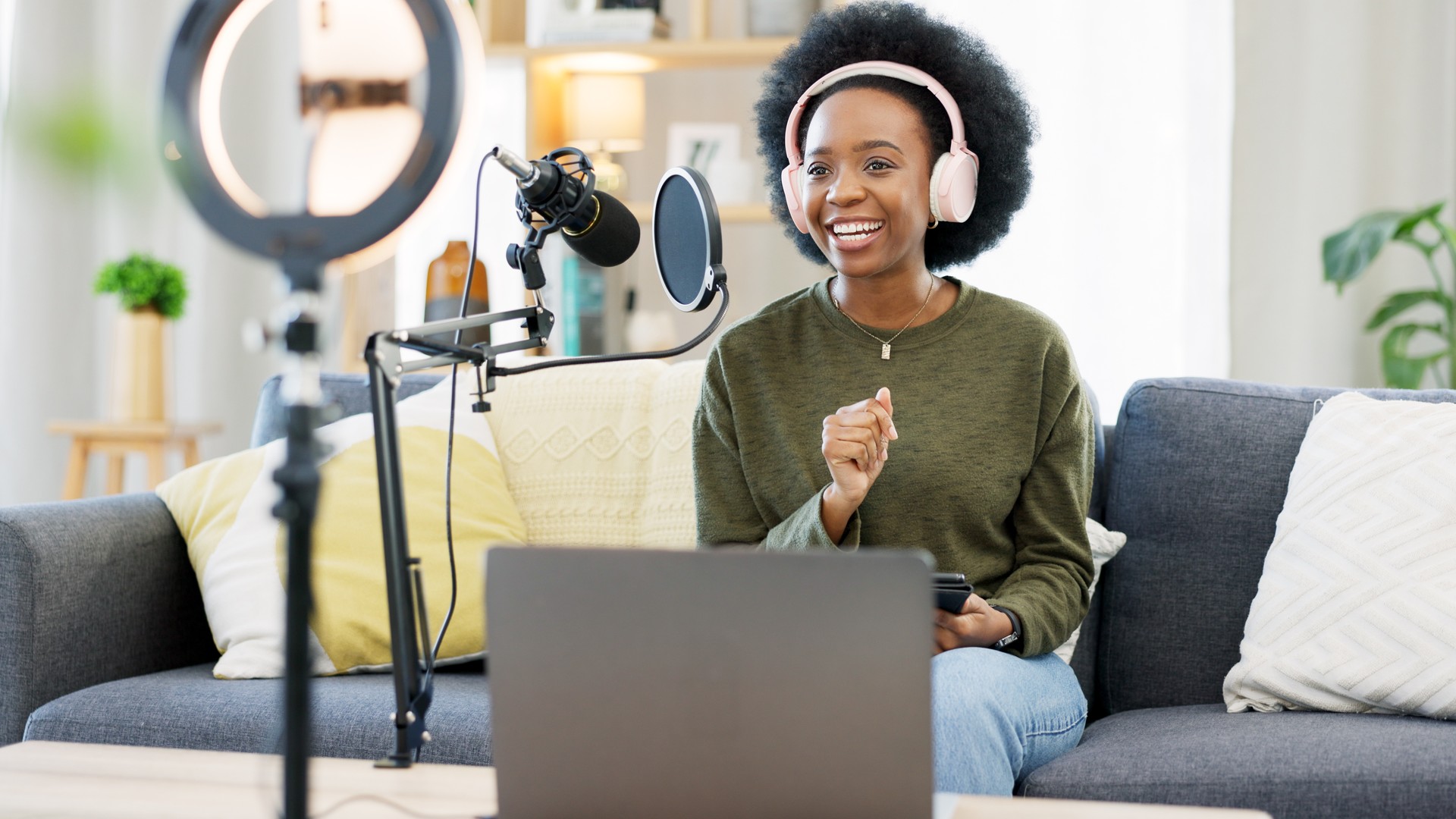 Headphones, laptop and black woman on microphone in podcast at home in living room of influencer. Technology, smile and content creator on mic for broadcast, communication and radio talk show on sofa